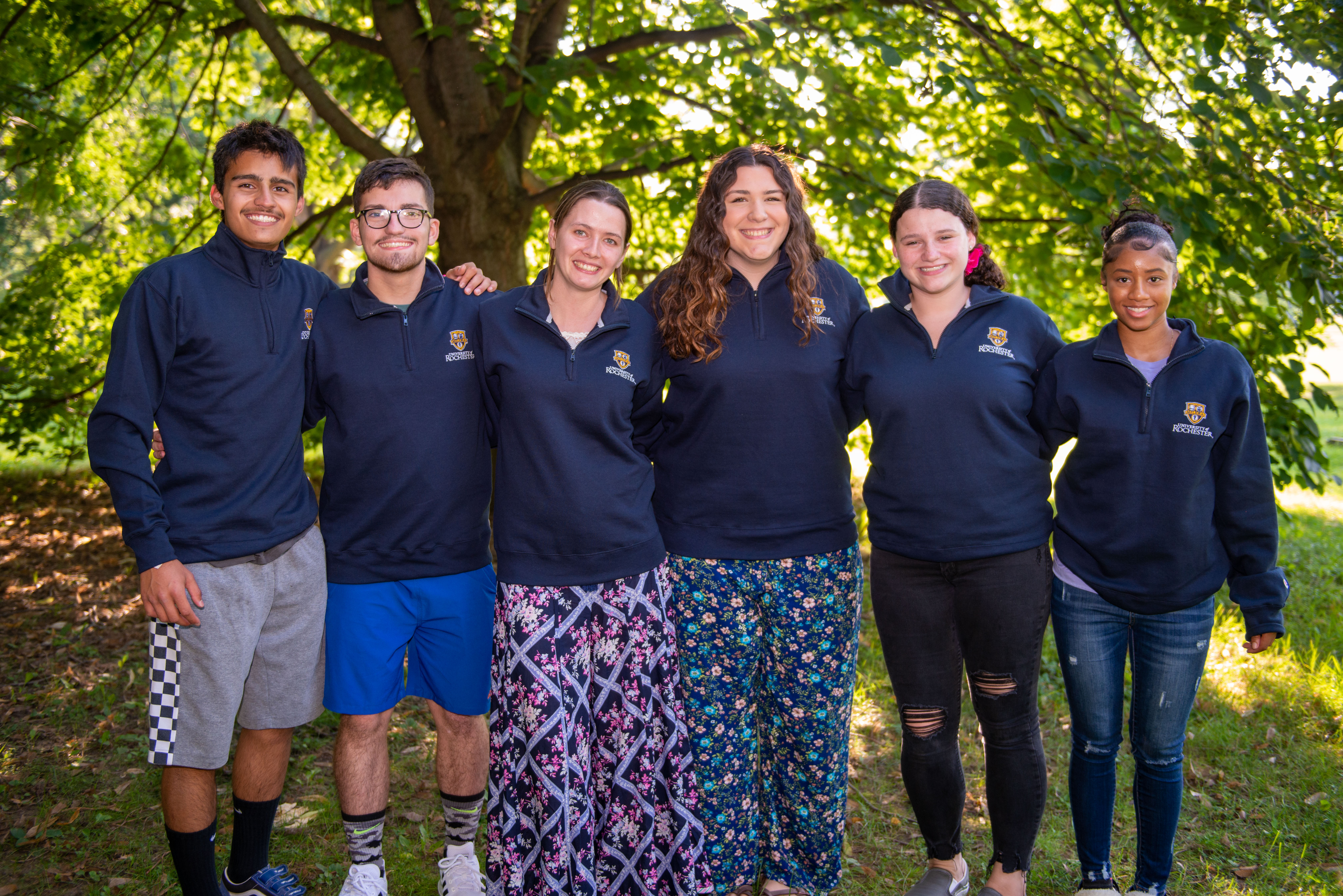 Six students wearing University of Rochester jackets stand together outdoors, smiling under a large tree.