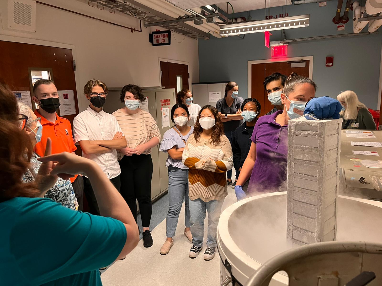 A group of masked students listens to an instructor in a University of Rochester lab, gathered around a large scientific instrument.