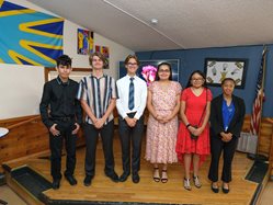 Six students in formal attire stand side by side in a decorated room with flags and artwork, smiling at the camera.