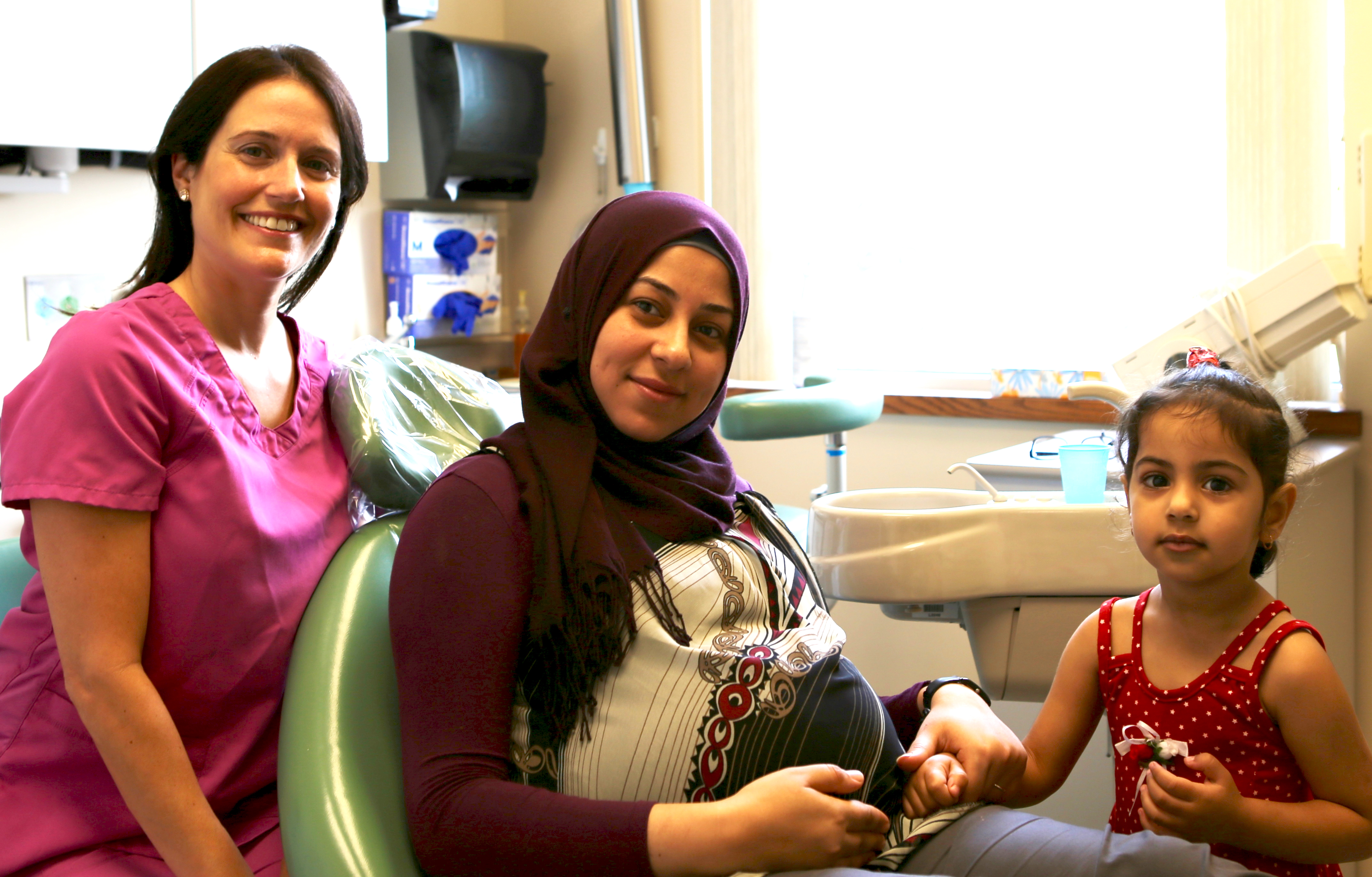 a pregnant woman sits in a dental chair with a child next to her. A dental professional sits nearby.