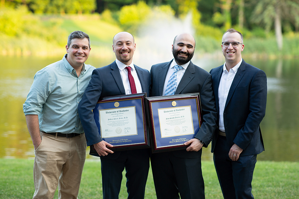 Four men in formal suits display certificates, posing by a calm lake on a sunny day.