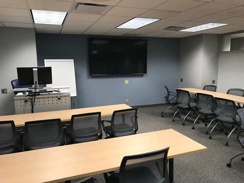 A classroom with tables and chairs, a teaching station with a computer, a large wall-mounted monitor, and a portable whiteboard.